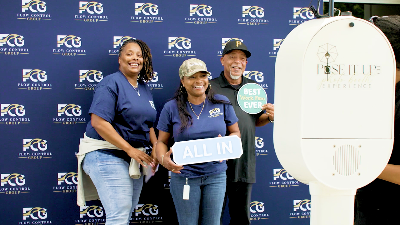 Three smiling people pose in front of a company-branded backdrop, holding signs that read “ALL IN.