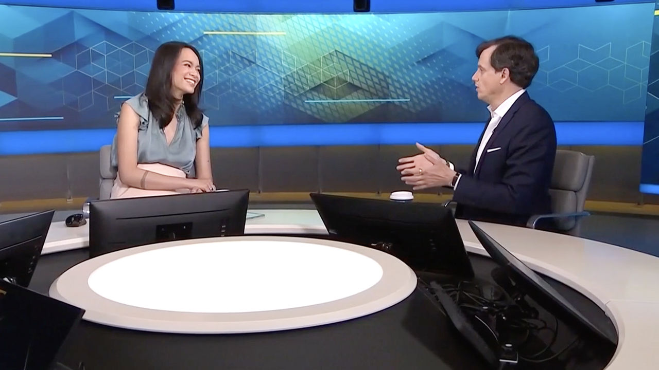 Two people seated at a news studio desk during an on-air interview.