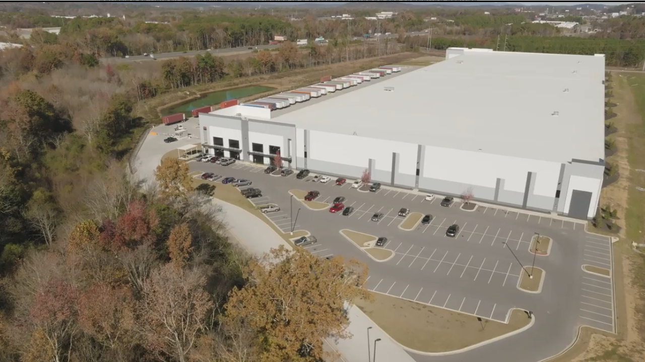 Aerial view of a large modern industrial warehouse and distribution facility with surrounding parking lots, trailers, and wooded landscape..